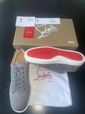 Gråa Christian Louboutin sneakers - Snygga gråa sneakers från Christian Louboutin i mocka med klassisk röd sula. Skorna har lågt skaft, rund tå och vita gummisulor. Kommer med originalkartong, dustbag och certifikat. Perfekta för dig som vill sticka ut med exklusiva detaljer.