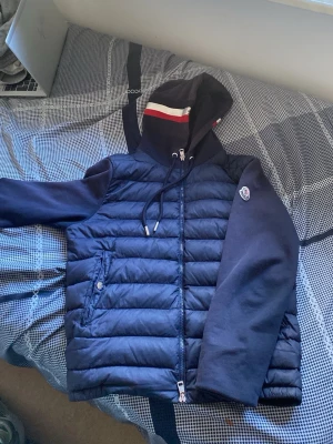 Mörkblå pufferjacka från Moncler - Snygg mörkblå pufferjacka från Moncler med vadderad front, mjuka ärmar och huva med röd och vit rand. Jackan har dragkedja, två fickor med knappar och Moncler-logga på ärmen. Perfekt för kalla dagar och riktigt trendig stil. Storlek S 