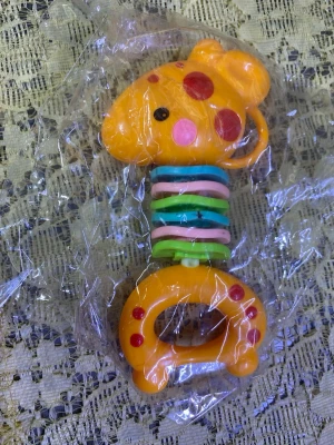 Färgglad skallra i form av giraff - En söt skallra i plast formad som en giraff med orange färg och röda prickar. Skallran har flera färgglada ringar i grönt, blått och rosa runt halsen. Perfekt för små barn att greppa och leka med.
