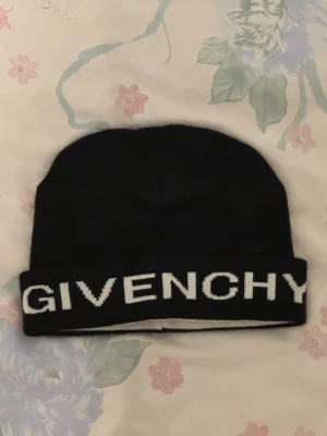 Svart mössa från Givenchy - Snygg svart mössa från Givenchy med vit logotyp runt kanten. Klassisk beanie-modell i mjukt stickat material, perfekt för att hålla värmen och ge en trendig look. Passar dig som gillar exklusiva accessoarer och streetstyle.