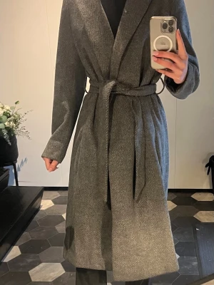 Grå lång kappa med knytskärp - Stilren grå kappa i klassiskt fiskbensmönster med bred krage och knytskärp i midjan. Lång modell som ger en snygg siluett, perfekt för kyliga dagar. Tidlös design som passar till många outfits.