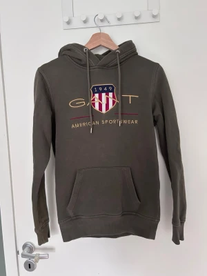 Olivgrön hoodie från Gant - Snygg olivgrön hoodie från Gant med broderad logga och texten 'American Sportswear' på bröstet. Klassisk känguruficka framtill, justerbar huva med dragsko och ribbade muddar. Perfekt för en avslappnad och sportig stil.