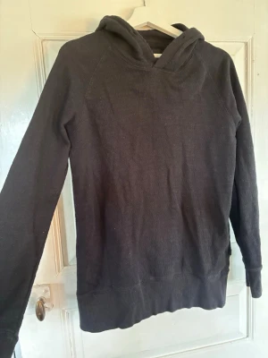 Svart hoodie från Tiger of Sweden - Mysig svart hoodie från Tiger of Sweden Jeans i storlek S. Tillverkad i ett mjukt stickat material med huva och långa ärmar. Enkel och stilren design utan tryck eller dragkedja, perfekt för en avslappnad look. Hoodien saknar tyvärr snöret