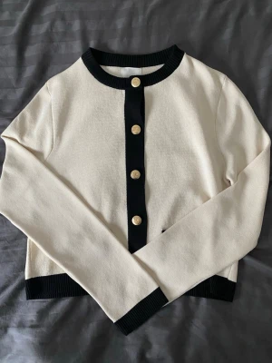 Vit kofta med svarta detaljer H&M - Stilren vit kofta från H&M med svarta ribbade kanter och guldfärgade knappar framtill. Koftan har rund halsringning och långa ärmar, perfekt för en chic och enkel look. Materialet är mjukt och bekvämt, passar till många outfits.