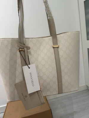 Beige mönstrad handväska Anna Field - Stilren handväska från Anna Field i beige med diskret mönster och ljusbeiga detaljer. Väskan har två långa axelband, guldfärgade metalldetaljer och medföljande matchande korthållare. Tillverkad i konstläder med elegant finish.