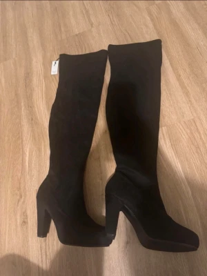 Svarta overknee stövlar med klack - Snygga svarta overknee stövlar med hög klack och rundad tå. Stövlarna är i mockaimitation och har en stilren, slimmad passform som går över knäet. Perfekta för att lyfta din outfit med en trendig touch.