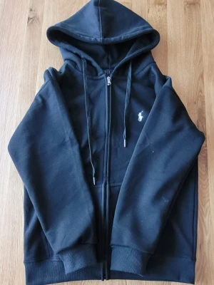 Svart Ralph Lauren - Oanvänd svart hoodie med dragkedja från Polo Ralph Lauren. Klassisk design med huva och dragsko, samt vit broderad logga på bröstet. Mjuk bomullsblandning och ribbade muddar vid ärmslut och nederkant. Perfekt för en avslappnad och stilren look. 