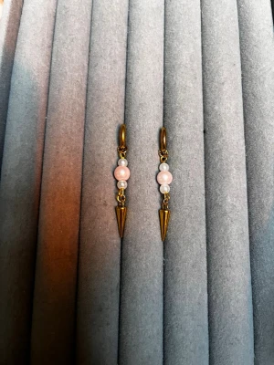 Guldörhängen med rosa pärlor och spikes - Snygga örhängen i guldfärgad metall med två ljusrosa pärlor och en liten vit pärla på varje. Hängande design med spetsig spike-detalj längst ner. Perfekta för att ge din look en cool och trendig touch.