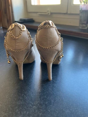 Beige pumps med guldkedjor från Moda Pelle - Snygga beige pumps från Moda Pelle med spetsig tå och höga klackar. Skorna är dekorerade med flera guldfärgade kedjor och små hängen som ger en unik och trendig look. Tillverkade i mockaimitation för en lyxig känsla.