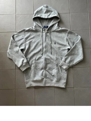 Grå zip hoodie från Polo Ralph Lauren - Snygg grå hoodie med dragkedja från Polo Ralph Lauren. Klassisk design med huva och dragsnören, två fickor framtill och den ikoniska lilla loggan broderad på bröstet. Tillverkad i mjuk bomullsblandning som känns skön mot huden.