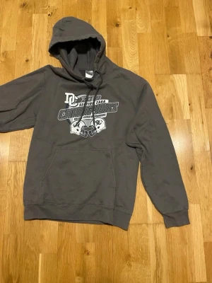 Grå hoodie med tryck Champions stl S - Säljer en grå hoodie från DC med stort tryck på bröstet där det står 'Champions' och motiv av två amerikanska fotbollshjälmar. Hoodien har huva med dragsko och en stor magficka. Perfekt för dig som gillar sportig stil och vill ha något bekvämt och snyggt.