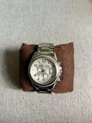 Silverfärgad klocka Michael Kors - Snygg silverfärgad klocka från Michael Kors med länkarmband i metall och glittrande stenar runt urtavlan. Inställd på ca 16-17 cm runt handleden men går såklart att ändra på. Klockan passar både kvinnor och män och är en extremt fin klocka. Bara att skicka om du undrar något och pris kan diskuteras vid snabb affär.