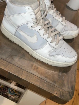 Nike vita höga sneakers - Snygga höga sneakers från Nike i vitt och ljusgrått läder. Klassisk design med perforerad tå, vadderad plös och Nike swoosh på sidorna. Skorna har vita snören och en robust yttersula som ger bra grepp. Perfekta för dig som gillar streetwear och sportig stil.