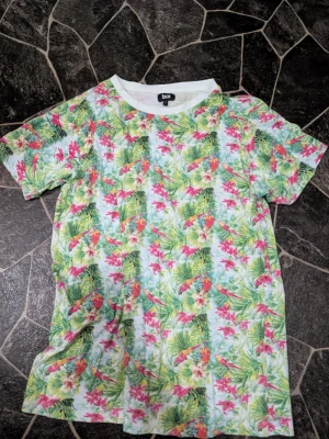 Blommig t-shirt från Izco stl L - Färgglad t-shirt från Izco med tropiskt blommönster i grönt, rosa och gult. T-shirten har vit krage och korta ärmar, perfekt för en somrig vibe. Materialet känns mjukt och lätt, troligtvis bomull eller bomullsblandning.