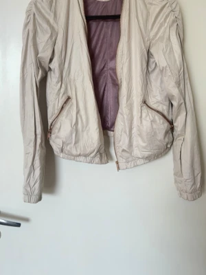 Beige vindjacka med dragkedjor - Stilren beige vår/sommar jacka med två snedställda fickor med dragkedja och lätt puffade axlar. Jackan har resår i nederkant och ärmar samt ett tunt, glansigt innerfoder i ljuslila. Perfekt för blåsiga dagar och enkel att matcha till olika outfits.