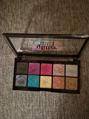 Glittrig ögonskuggspalett från NYX - Snygg ögonskuggspalett från NYX med 10 glittriga nyanser. Färgerna inkluderar svart, silver, turkos, guld, rosa, röd, koppar, brons och champagne. Paletten har fyrkantiga skuggor i en svart plastförpackning med genomskinligt lock. Swatshad i några/någon skugga. 