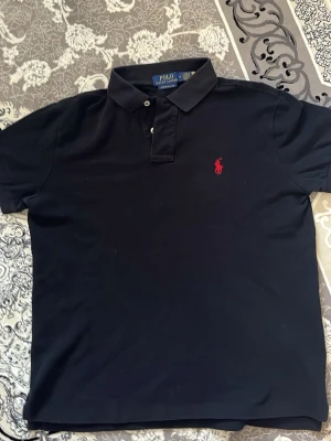 Svart pikétröja från Polo Ralph Lauren - Svart pikétröja från Polo Ralph Lauren med röd broderad logga på bröstet. Tröjan har klassisk krage, knappar framtill och är i en slim passform. Perfekt för en stilren och enkel look. Har bara använt en gång!