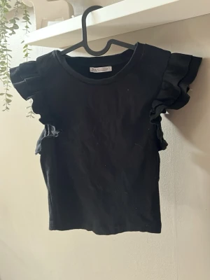 Svart volangtopp från Zara - Svart topp från Zara med volangdetaljer på axlarna. Modellen är croppad och har en tight passform. Perfekt att styla med jeans eller kjol för en trendig look.