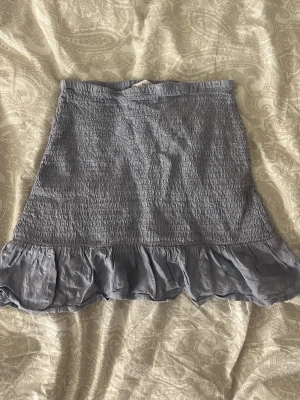 Blå volangkjol från MNG - Supersöt blå kjol från MNG med smockad midja och volangkant nedtill. Kjolen har en kort och luftig modell som passar perfekt till varma dagar. Enkel att matcha med både toppar och sneakers för en chill look. Ny 