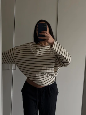 Randig croppad långärmad topp - Vit croppad långärmad topp med gröna ränder och loose passform. Tillverkad i mjuk bomull och har klassisk rund hals. Perfekt för en chill och avslappnad look.