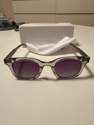 Grå CHIMI solglasögon  - Snygga solglasögon från CHIMI med transparent grå båge ljuset gör stt glaset ser lite lila ut. Modellen är klassisk med raka linjer och markerad ram. Perfekt accessoar för att lyfta din stil och skydda ögonen mot solen. Kommer med originalfodral och putsduk.