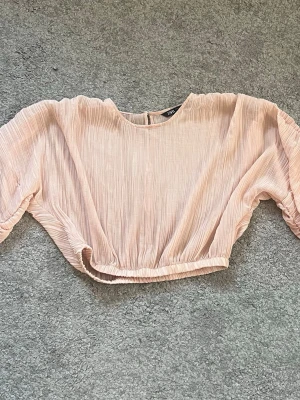 Ljusrosa croppad topp från Zara - Säljer en ljusrosa croppad topp från Zara med ballongärmar och resår i midjan. Toppen har ett skirt, plisserat tyg som ger en luftig känsla och rund halsringning. Perfekt för dig som gillar trendiga och feminina plagg. Använd 2 gånger 