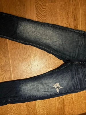 Mörkblå slitna jeans från Replay - Säljer ett par mörkblå jeans från Replay med slitningar och en trasig detalj på ena benet. Jeansen har en klassisk femficksmodell och tydliga tvätteffekter för en cool, använd look. Skriv för frågor.