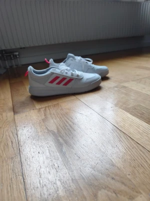 Vita Adidas sneakers med rosa detaljer - Snygga vita sneakers från Adidas med klassiska rosa ränder på sidan och matchande rosa detaljer på häl och plös. Skorna har lågt skaft, vita snören och ovandel i syntetmaterial. Perfekta för dig som vill ha en clean och sportig look.