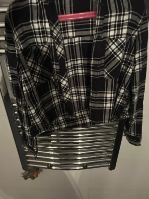 Svartvit rutig skjorta från Lindex - Klassisk svartvit rutig skjorta från Lindex med två bröstfickor och knappar framtill. Skjortan har lång ärm och är tillverkad i mjukt bomullstyg. Perfekt för en avslappnad och cool stil.