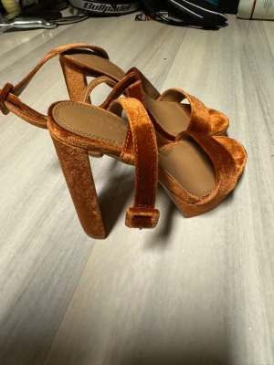 Orange sammetsandaler med klack - Säljer ett par trendiga sandaler i orange sammet med breda remmar och kraftig blockklack. Sandalerna har öppen tå och justerbart spänne runt ankeln. Perfekta för dig som vill sticka ut med en färgstark och cool look.