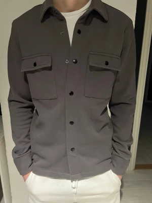Mörkgrå overshirt med fickor - Stilren mörkgrå overshirt med två stora bröstfickor och svarta tryckknappar framtill. Overshirten har klassisk krage och raka ärmar med knapp vid ärmslut. Materialet ser ut att vara mjukt och något stretchigt, perfekt för lager-på-lager. Passar till många olika stilar.
