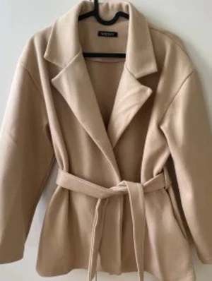 Beige kappa från missay - Stilren beige kappa från missäy med knytskärp i midjan. Kappan har en rak passform och är tillverkad i ett mjukt material som ger en snygg siluett. Perfekt för dig som vill ha en enkel men trendig look.