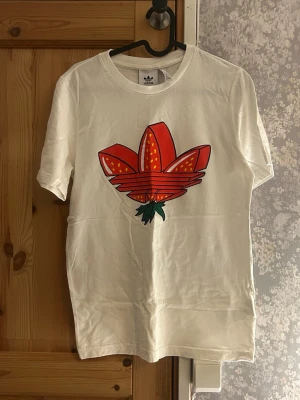 Vit Adidas t-shirt med jordgubbstryck - Vit t-shirt från Adidas med stort tryck av röda jordgubbar. Mycket bra skick aldrig använd. Hör av vid frågor!💗