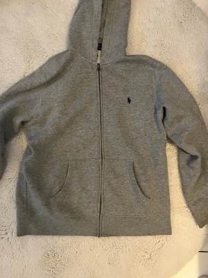 Grå hoodie från Polo Ralph Lauren - Snygg grå zip-hoodie från Polo Ralph Lauren med klassisk logga broderad på bröstet. Hoodien har dragkedja, två stora fickor fram och en mjuk insida. Perfekt för chill dagar och enkel att matcha med allt. Tillverkad i bomullsmix för skön känsla.