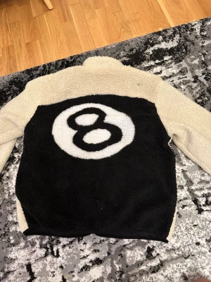Beige teddyjacka med 8-ball design - Säljer en beige teddyjacka med svart ryggparti och stor vit 8-ball design. Jackan har svart bröstficka, två sidofickor med dragkedja och hög krage. Materialet är mjukt och fluffigt, perfekt för kalla dagar. Stängs med dragkedja framtill.