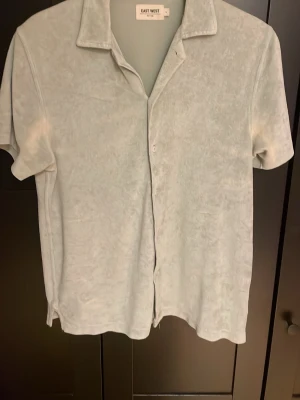 Ljusgrå kortärmad skjorta East West - Säljer en ljusgrå kortärmad skjorta från East West. Skjortan har klassisk krage, knappar framtill och är gjord i ett mjukt material som ger en avslappnad vibe. Perfekt till sommaren och enkel att styla med jeans eller shorts. Nästa aldrig använd..
