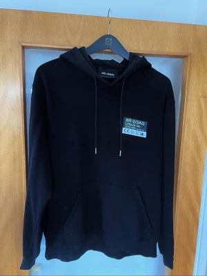 Svart hoodie Axel Arigato batteritryck - Svart hoodie från Axel Arigato med huva och dragsnören. Tryck på bröstet med texten 'BR-2/3AG Lithium Ion' och stor batterigrafik på ryggen med detaljer i grått och orange. Stor känguruficka framtill. Perfekt för dig som gillar streetwear och unika prints.