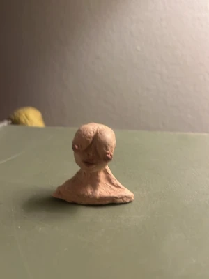 Liten handgjord lerfigur - Unik liten skulptur i beige lera, formad som ett huvud med stora ögon och enkel kropp. Perfekt som dekoration eller samlarobjekt för dig som gillar ovanliga konstverk.
