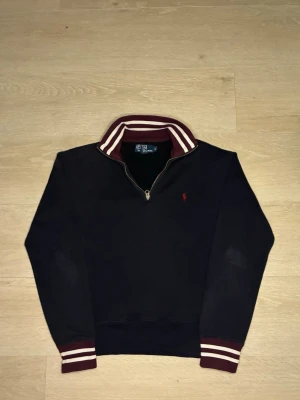 Svart stickad tröja Polo Ralph Lauren - Mörkblå stickad tröja från Polo Ralph Lauren med halv dragkedja och vinröd krage med vita ränder. Klassisk röd logga broderad på bröstet. Ribbstickade muddar och krage ger en sportig look. Perfekt till höst och vinter.