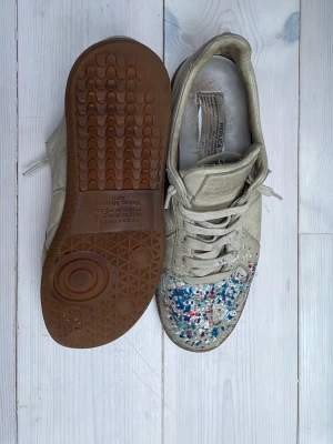 Maison Margiela gats - Tjena, säljer dessa feta maison margiela gatsen i en beige färg med blått, rött och vitt paint splatter. Perfekta skon för dig som vill ha ett par unika pjux till våren och sommaren. Skorna är i bra skick bortsätt från en liten defekt på insidan av hälen. Skorna är en size 40,5 men passar dig som bör size 41,5 och till och med 42. Det är en väldigt sällsynt och eftertraktad design och därav priset. Och skorna är självklart äkta. Om det skulle vara något du undrar så är det bara att höra av dig🕺