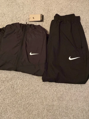 Svart Nike träningsset  - Säljer ett svart träningsset från Nike Running med jacka och byxor. Båda plaggen har vita Nike-loggor och är tillverkade i lätt, funktionellt material som passar perfekt för löpning eller annan träning. Jackan har dragkedja och byxorna har resår i midjan. Det är storlek M men den är inte så stor i storlek det är därför jag säljer eftersom den är lite tajt på mig. Helt oanvänd 