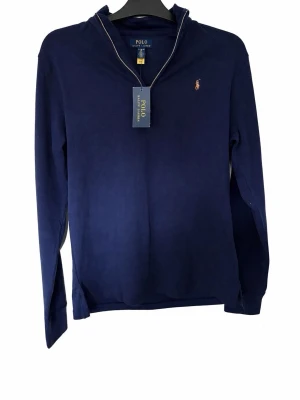 Marinblå tröja från Polo Ralph Lauren - Klassisk marinblå half zip tröja från Polo Ralph Lauren med liten broderad logga på bröstet. Tröjan har v-ringad hals med diskreta kontrastsömmar och är tillverkad i mjukt material. Perfekt för en stilren och avslappnad look. Nypris=1899kr