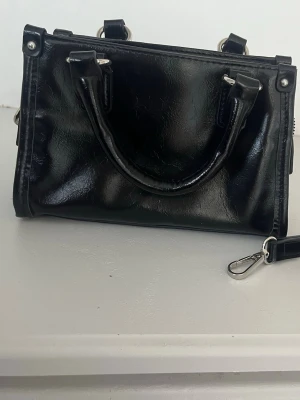 Svart handväska  - Snygg svart handväska i glansigt konstläder med silverfärgade metalldetaljer. Väskan har både handtag och avtagbar axelrem samt dragkedja upptill. Säljer då jag har 2 andra svarta🥰