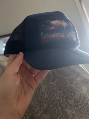 Marinblå truckerkeps från Natone - Snygg svart truckerkeps från Natone med mesh på sidorna och ett coolt tryck framtill som visar fyrverkerier över en stadssilhuett. Justerbar baktill och med böjd skärm. Perfekt för dig som vill ha en unik och stilren keps.