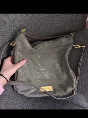 Grå axelväska från Marc by Marc Jacobs - Snygg grå axelväska i mjukt, strukturerat läder från Marc by Marc Jacobs. Väskan har guldfärgade metalldetaljer, justerbar axelrem och en metallbricka med logga framtill. Perfekt för dig som vill ha en stilren och rymlig väska. Skavanker syns på bilderna.