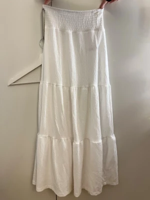 Vit långkjol från 157 - Luftig och flowy vit långkjol från 157 med smock i midjan och volangdetaljer. Kjolen har en enkel och stilren look, perfekt för varma dagar. Tillverkad i ett lätt och mjukt material som känns skönt mot huden.