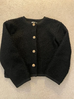 Svart kofta från Gina Tricot - svart kofta från Gina Tricot med rund halsringning och dekorativa guldfärgade knappar framtill. Koftan har lång ärm och är gjord i ett mjukt material som ger en trendig look. Perfekt för en stilren look.