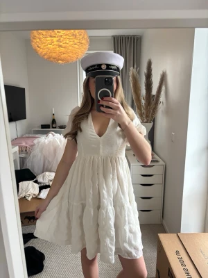 Vit kort klänning med v-ringning - Söt vit kortklänning med v-ringning, puffärmar och rosett i ryggen. Klänningen har en volangig kjol och är tillverkad i ett lätt, strukturerat tyg som ger en luftig känsla. Perfekt för student eller sommarens tillfällen. Från bubbelroom, endast använd 1 gång därav i nyskick. Storlek xxs men passar mig som bär xs också.