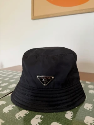 Svart bucket hat från Prada - Snygg svart bucket hat från Prada med den ikoniska metalltriangeln framtill. Hatten är tillverkad i slitstark nylon och har en klassisk rund form med bred, nedåtlutande brätte. Perfekt accessoar för en trendig och avslappnad stil.