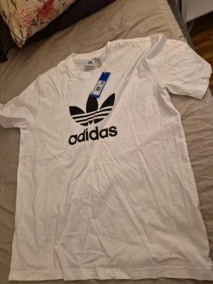 Vit Adidas t-shirt med stor logga - Snygg vit t-shirt från Adidas med den klassiska svarta Trefoil-loggan tryckt framtill. T-shirten har rund halsringning och korta ärmar. Tillverkad i mjuk bomull som känns skön mot huden. Perfekt för en avslappnad och sportig stil.